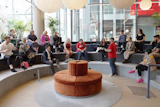 A group of people gathered in a modern, open space, attentively listening to a speaker in a red shirt, with a videographer recording the session.